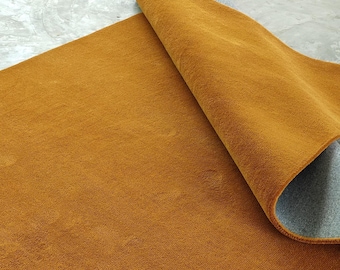 May include: A rectangular, mustard-colored rug with a rolled-up edge, revealing a gray backing. The rug has a soft, textured surface, and is placed on a concrete floor. The color is a warm, earthy tone.