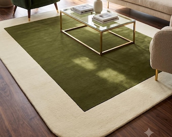 Puede incluir: Una alfombra rectangular que presenta un gran panel central de color verde oliva sólido, enmarcado por un borde ancho de color crema con esquinas redondeadas. La alfombra se sitúa sobre un suelo de madera oscura bajo una mesa de centro de cristal.