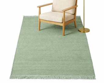 May include: A light green rectangular rug with fringe, a light wood armchair with a cream cushion, and a gold floor lamp with a white lampshade. The rug is the focal point of the image.