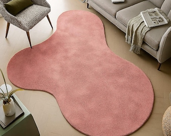 May include: A large, irregularly shaped rug in a soft, muted rose color. The rug has a plush texture and covers a significant portion of the room's floor. The design is modern and organic, adding a touch of warmth and style to the space.