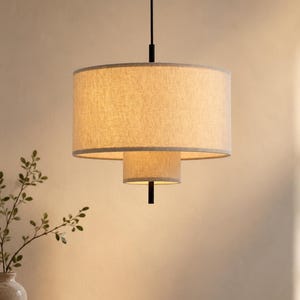 Może przedstawiać: Dwupoziomowa lampa wisząca z dwoma cylindrycznymi abażurami z tkaniny w kolorze jasnego beżu. Klosze są ułożone jeden na drugim, z większym górnym i mniejszym dolnym bębnem, zawieszone na cienkim czarnym metalowym pręcie i czarnym przewodzie.