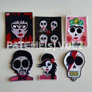 May include: A collection of embroidered patches featuring stylized skull designs. The patches include portraits of iconic figures, each with unique details and vibrant colors. The text "PATCHE(S) Vol. 2" is displayed across the patches.