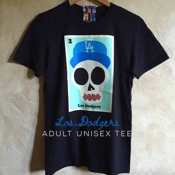 Puede incluir: Camiseta unisex negra para adultos con un gráfico de una calavera con una gorra de béisbol azul con la letra "A". El gráfico está sobre un fondo azul claro con las palabras "Los Dodgers" y "Adult Unisex Tee" en azul.