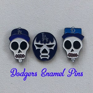 May include: Three enamel pins featuring a skull design with a baseball cap, a wrestling mask, and a baseball cap with the letter "D". The pins are white, blue, and black. The text "Dodgers Enamel Pins" is written in blue cursive.