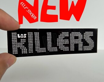 Peut inclure: Un autocollant rectangulaire noir mesurant 10,16 par 3,05 cm avec le texte LOS KILLERS. Les lettres sont composées d'un motif répétitif de petites icônes de crânes blancs. L'autocollant a une finition lisse et un design contrasté.