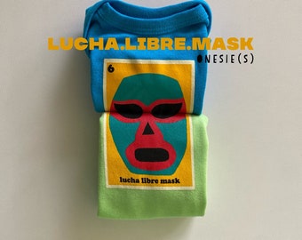 Peut inclure: Deux bodies pliés, un bleu et un vert, superposés. Chacun présente un motif de masque de lucha libre. Le texte "LUCHA.LIBRE.MASK ONESIE(S)" est imprimé en jaune. Vêtements pour bébés.