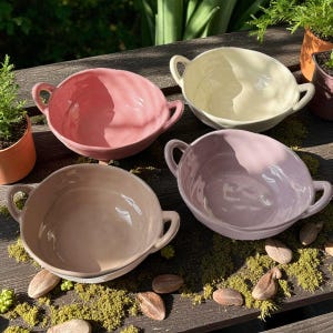 Peut inclure: Quatre bols en céramique rose, beige et lavande, chacun avec deux petites anses. Les bols sont disposés sur une surface en bois avec de petites plantes et de la mousse en arrière-plan. Les bols mesurent environ 15 cm de diamètre.
