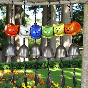 Puede incluir: Seis campanillas de viento con forma de gato de colores colgando de una rama de árbol. Cada gato de cerámica es de un color diferente, incluyendo naranja, blanco, azul, verde, amarillo y naranja. Debajo de cada gato hay una campana plateada y un badajo en forma de hoja.