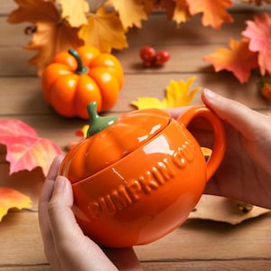 Pode incluir: Uma caneca de cerâmica laranja em forma de abóbora com uma haste verde e as palavras "PUMPKIN CUP" em relevo na lateral. A caneca é segurada nas mãos, com uma abóbora e folhas de outono ao fundo.