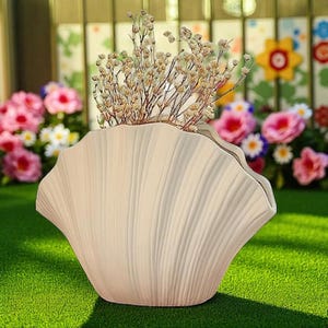 Może przedstawiać: Kremowy wazon w kształcie muszli z pionowymi żłobieniami. Wazon zawiera suszone, jasno brązowe kwiaty. Tło przedstawia zielony trawnik i kolorowe kwiaty, sugerując otoczenie ogrodu.