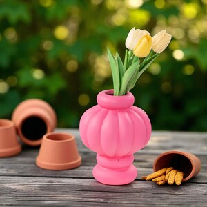 Peut inclure: Un vase rose vif, de forme bombée, contenant deux tulipes jaunes et blanches. Plusieurs petits pots en terre cuite sont disposés autour du vase sur une surface en bois, avec un arrière-plan vert flou.