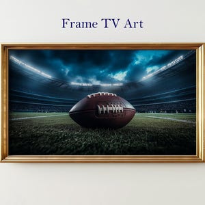 May include: A framed print featuring a football on a green field in a stadium. The football is brown with white laces. The stadium is lit with bright lights and a dark, cloudy sky. The text "Frame TV Art" is at the top.