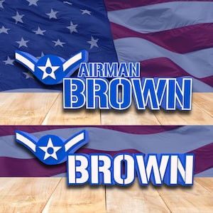 May include: A patriotic design featuring the text "AIRMAN BROWN" in bold blue letters with a white outline, accompanied by a blue emblem with a star and wings. The background includes the American flag and a wooden surface.