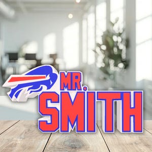 May include: A sign featuring the Buffalo Bills logo and the name "MR. SMITH" in bold, red letters with a blue outline. The logo is a white, red, and blue buffalo head. The sign is on a wooden surface.