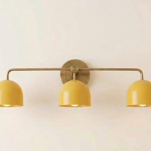 May include: A wall-mounted light fitting featuring a horizontal brass bar and a circular brass backplate. Three dome-shaped shades in a matte mustard yellow finish are attached to the bar, each housing a light bulb pointing downwards.