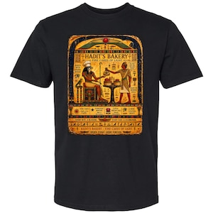May include: A black t-shirt featuring a graphic print with an Egyptian-themed bakery design. The artwork displays figures in ancient attire, cakes, and hieroglyphic-style text that reads Hadit's Bakery, Fine Cakes of Light, and other phrases.