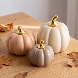 May include: Three ceramic pumpkin figurines in varying sizes and muted tones of cream, beige, and taupe. Each pumpkin features a smooth, glossy finish and a distinct, reflective gold-coloured stem at the top, creating a decorative set.