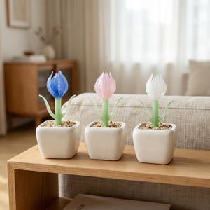 Op de afbeelding: Drie glazen tulpenbeelden in witte vierkante potten. Eén tulp is blauw, één is roze en één is wit. De stelen zijn groen en de potten zijn gevuld met kleine kiezels. De sculpturen worden tentoongesteld op een houten plank.