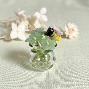Puede incluir: Un jarrón miniatura de vidrio transparente con lunares verdes que contiene varias flores de vidrio verde claro con centros amarillos y una pequeña figura de abeja de vidrio con rayas negras y amarillas y alas rosa claro sobre el arreglo floral.