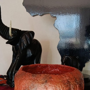 Puede incluir: Un cuenco redondo y texturizado de color marrón rojizo con un acabado exterior rugoso se sitúa en primer plano. Detrás, una figura de elefante de madera tallada en negro con un colmillo blanco y una silueta negra brillante del continente africano.