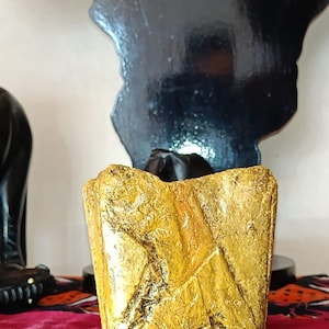 Peut inclure: Un petit récipient de forme carrée avec une surface texturée jaune doré. L'extérieur présente une finition rugueuse et inégale avec des indentations angulaires profondes et un aspect patiné, créant un aspect brut sur toutes les faces visibles.
