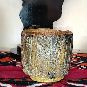 Peut inclure: Un pot de fleurs cylindrique en céramique avec un extérieur rugueux et texturé, présentant une finition gris anthracite et or métallique. Le bord est épais et irrégulier, tandis que la base est une bande solide de peinture dorée vive.
