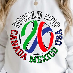 Może przedstawiać: Biała bluza z kolorową grafiką na Mistrzostwa Świata 2026. Projekt zawiera napis "WORLD CUP USA CANADA MEXICO" wok&oacute;ł stylizowanej liczby "26" w kolorach zielonym, czerwonym i niebieskim. Casualowy top z długim rękawem.