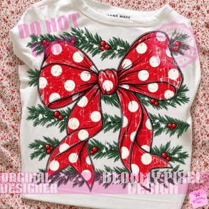 May include: A white long-sleeve shirt featuring a large red bow with white polka dots centered over a background of green pine branches with small red berries. The design has a distressed texture and the text HAND MADE is at the neckline.