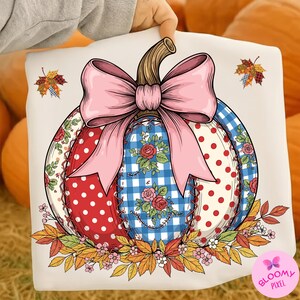 Pode incluir: Uma almofada decorativa com um design de abóbora em patchwork. A abóbora tem secções de bolinhas vermelhas, xadrez azul e padrões florais, com um grande laço rosa e um caule. Folhas de outono e flores rodeiam a base.