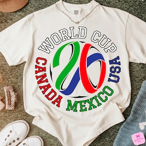 Może przedstawiać: Kremowa koszulka z okrągłą grafiką na Mistrzostwa Świata 26, z napisem "World Cup Canada USA Mexico" w kolorze czerwonym, zielonym i niebieskim. Numer "26" jest w kolorze zielonym, czerwonym i niebieskim. Koszulka leży na zielonej powierzchni.
