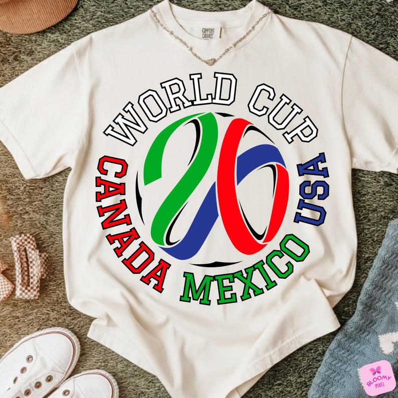 Może przedstawiać: Kremowa koszulka z okrągłą grafiką na Mistrzostwa Świata 26, z napisem "World Cup Canada USA Mexico" w kolorze czerwonym, zielonym i niebieskim. Numer "26" jest w kolorze zielonym, czerwonym i niebieskim. Koszulka leży na zielonej powierzchni.
