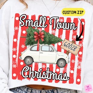 Puede incluir: Una sudadera blanca con un gráfico de un coche blanco antiguo que lleva un árbol de Navidad verde con un lazo rojo en el techo. El diseño incluye rayas verticales rojas y blancas, copos de nieve blancos y el texto Small Town Christmas.