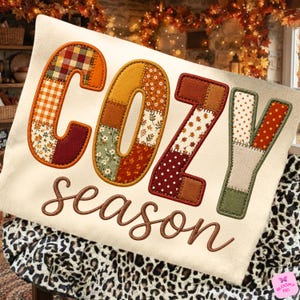 Könnte beinhalten: Ein cremefarbenes Sweatshirt mit dem Wort COZY in großen, gemusterten Stoffbuchstaben mit herbstlichen Drucken wie Karos, Blumen und Punkten. Darunter ist das Wort season in einer braunen, geschwungenen Schreibschrift aufgestickt.