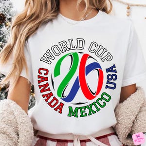 Może przedstawiać: Biała koszulka z kolorową grafiką na Mistrzostwa Świata 2026. Projekt zawiera napis "World Cup USA Canada Mexico" oraz piłkę nożną z numerem "26" w kolorach zielonym, czerwonym i niebieskim.