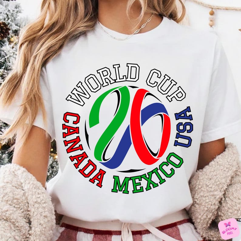 Może przedstawiać: Biała koszulka z kolorową grafiką na Mistrzostwa Świata 2026. Projekt zawiera napis "World Cup USA Canada Mexico" oraz piłkę nożną z numerem "26" w kolorach zielonym, czerwonym i niebieskim.