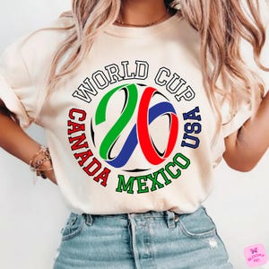 Może przedstawiać: Kremowa koszulka z kolorową grafiką na Mistrzostwa Świata USA, Kanada i Meksyk. Projekt zawiera napis "WORLD CUP" oraz cyfry "26" w kolorze zielonym, czerwonym i niebieskim, otoczone nazwami kraj&oacute;w.