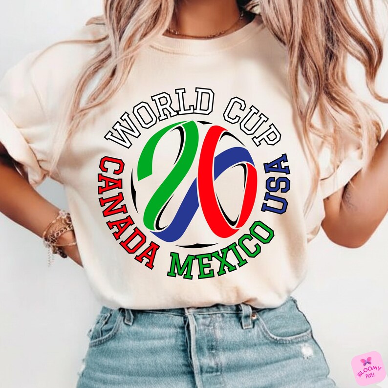 Może przedstawiać: Kremowa koszulka z kolorową grafiką na Mistrzostwa Świata USA, Kanada i Meksyk. Projekt zawiera napis "WORLD CUP" oraz cyfry "26" w kolorze zielonym, czerwonym i niebieskim, otoczone nazwami kraj&oacute;w.