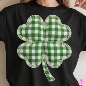 May include: Black t-shirt featuring a large, four-leaf clover design. The clover is made of green and white checkered fabric, with a cream-colored stitched border. The design is centered on the shirt.