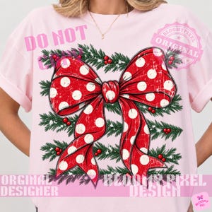 May include: A light pink t-shirt featuring a large red bow with white polka dots, layered over a background of green pine branches with small red berries. The design has a distressed, vintage texture throughout the bow and the foliage.