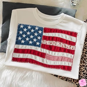 Puede incluir: Camiseta blanca con un diseño de bandera estadounidense cosido. La bandera tiene un campo azul con estrellas blancas y rayas rojas y blancas. Las rayas incorporan patrones florales y de cuadros.