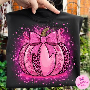 Könnte beinhalten: Schwarzes Sweatshirt mit einem rosa Kürbisdesign, einer großen Schleife und rosa Bändern. Der Kürbis hat ein Leopardenmuster und rosa Spritzer. Das Design enthält den Text "BLOOMY PIXEL".