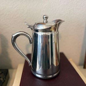 May include: A polished silver-tone metal pitcher with a hinged lid, a small spherical finial, a curved handle, and a wide pouring spout. The pitcher features a smooth, reflective surface and a cylindrical body with a slightly tapered neck.