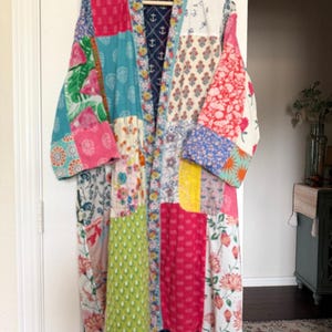 May include: A long-sleeved patchwork robe featuring a variety of colourful floral, geometric, and botanical fabric patterns in shades of pink, yellow, green, and blue. The interior lining displays a dark blue fabric with a white anchor print.