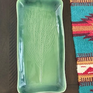 May include: A rectangular ceramic tray with a glossy green glaze and a debossed fern leaf pattern centered on the surface. The tray features slightly raised, curved edges and a smooth, reflective finish that highlights the botanical design.