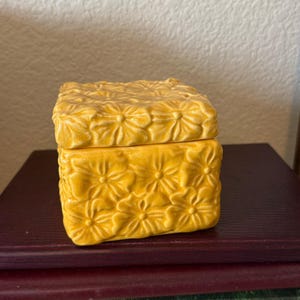 May include: A square yellow ceramic box with a lid, featuring a raised, embossed floral pattern covering all visible sides and the top surface. The glaze has a glossy finish with subtle variations in the yellow color tone across the texture.