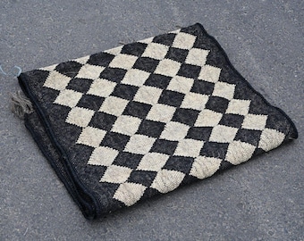 May include: A square, woven textile with a black and off-white checkerboard pattern. The edges are finished with a dark border and fringe. The textile is lying on a gray surface.