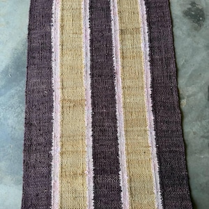 May include: A rectangular woven rug with vertical stripes. Alternating stripes of dark brown, tan, and light pink create a textured pattern. The rug appears to be made of natural fibers, suitable for a rustic or bohemian decor.