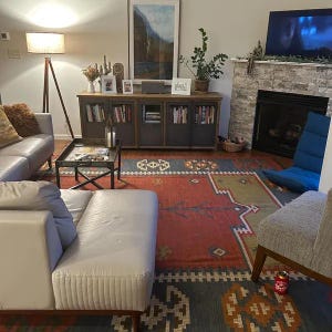 May include: A living room scene featuring a gray leather sofa, a patterned rug with red and blue hues, and a wooden cabinet with glass-fronted doors filled with books. A floor lamp and a fireplace add to the cozy ambiance.