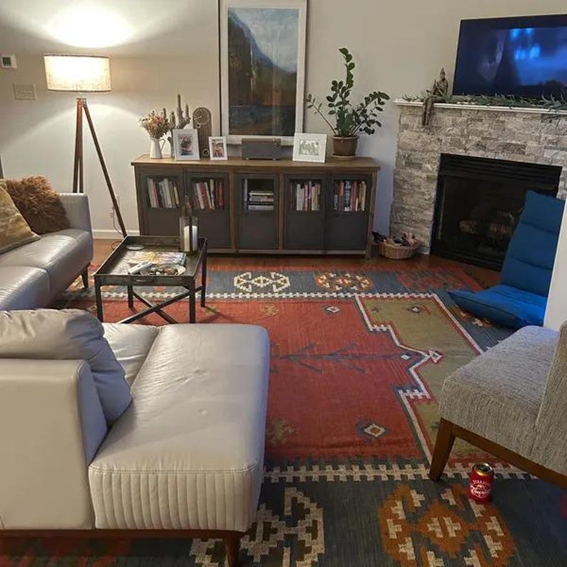 May include: A living room scene featuring a gray leather sofa, a patterned rug with red and blue hues, and a wooden cabinet with glass-fronted doors filled with books. A floor lamp and a fireplace add to the cozy ambiance.