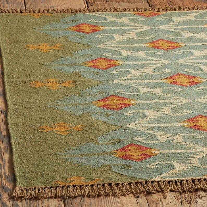 May include: A rectangular rug with a geometric pattern. The rug features a green border and a central design in shades of blue, cream, orange, and yellow. The rug has a fringed edge.